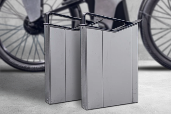 Two Rectangular, Metal Bike Battery Cases Are Positioned Side By Side On A Concrete Surface With A Blurred Bicycle Wheel In The Background, Highlighting The Cutting-edge Technology Behind Electric Scooters Rental In The UK.